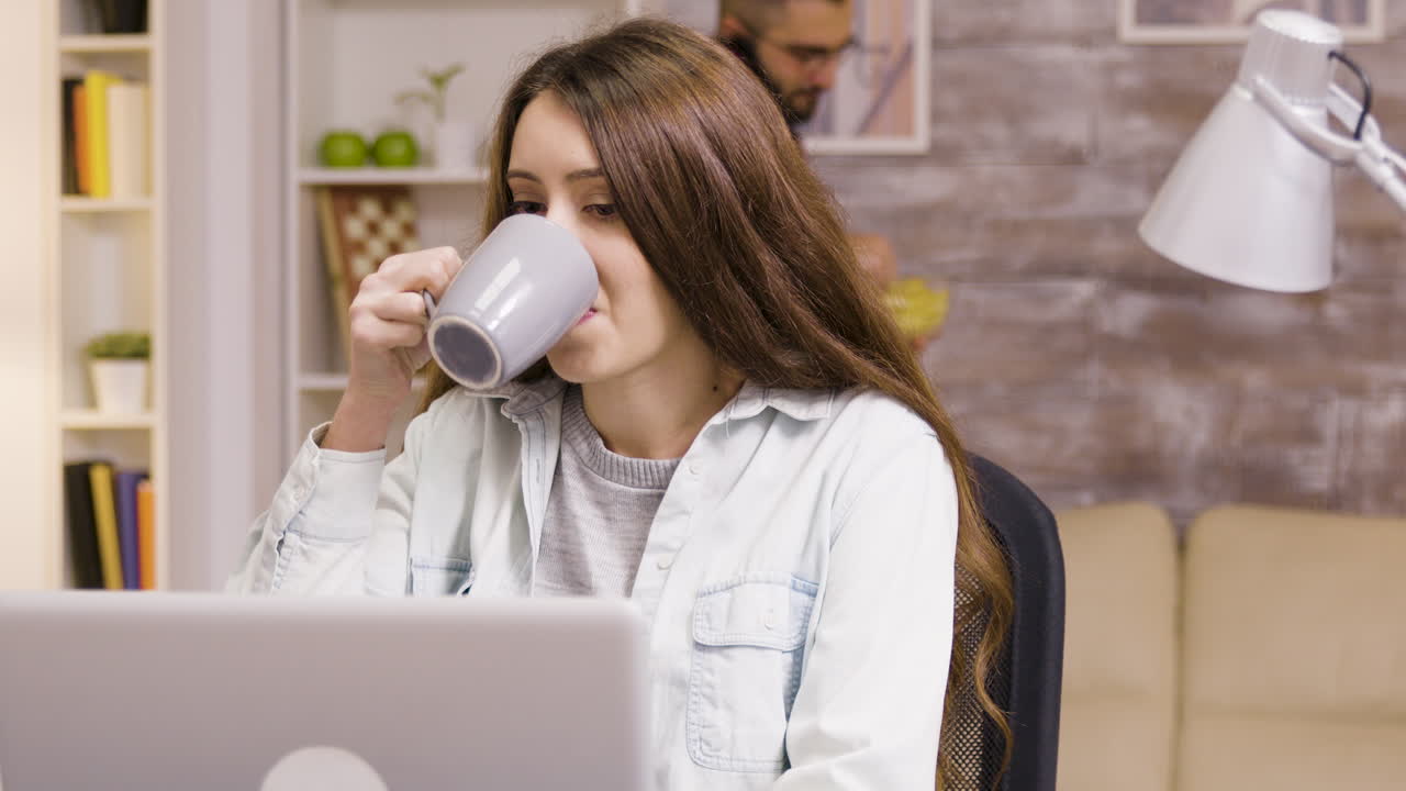 Woman working at her laptop with a man eating in the background
