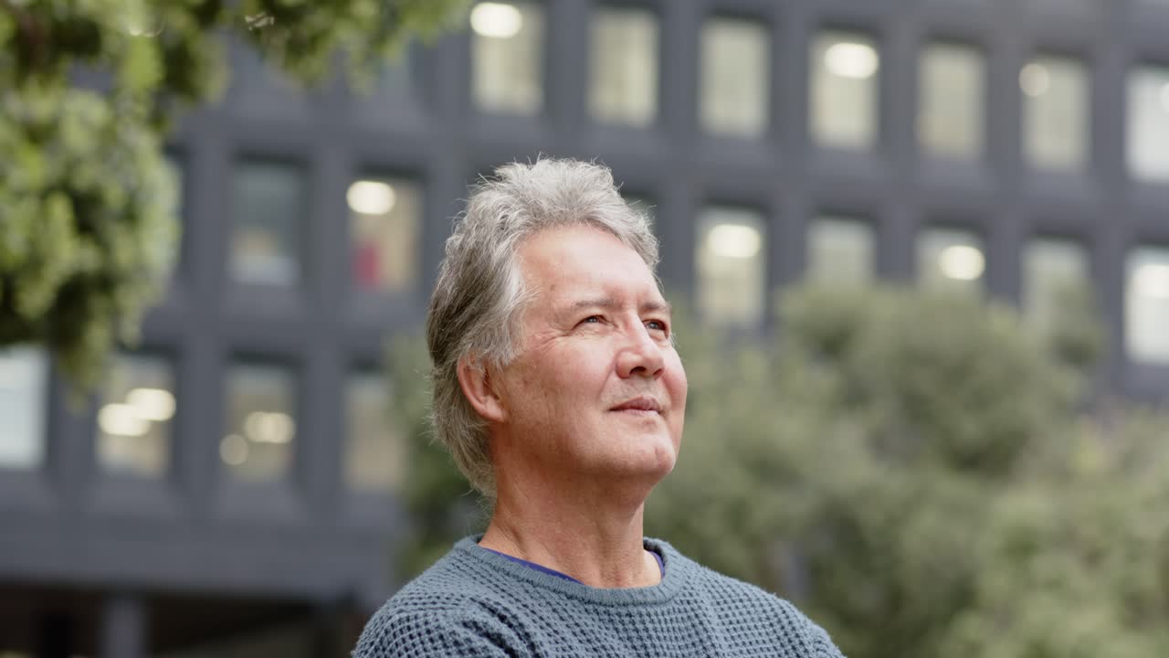 一位老年男士在辦公廣場抬頭望，轉向鏡頭，自信地抱著雙臂並微笑。