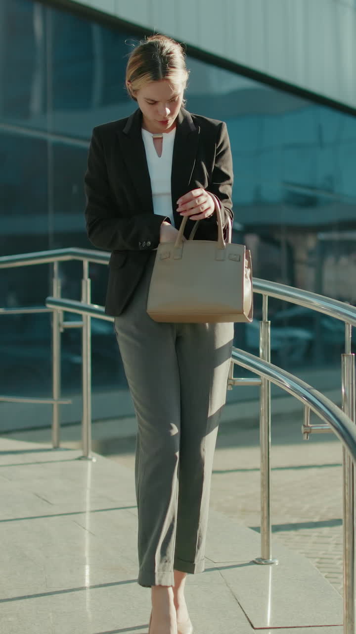 Professional businesswoman leaves work, walking down ramp with metal railings, checking handbag thoughtfully, retrieving keys, modern glass building and parked cars reflecting urban office setting