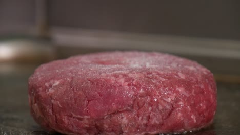 close-up of a raw burger patty on a restaurant grill as black pepper and salt are ground on top