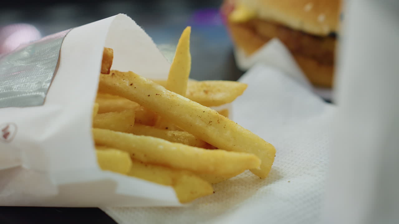 Close up of dark skinned finger reaching for crispy golden seasoned fries on white napkin beside sesame burger during lunch in indoor fast food setting under soft daylight with blurred background