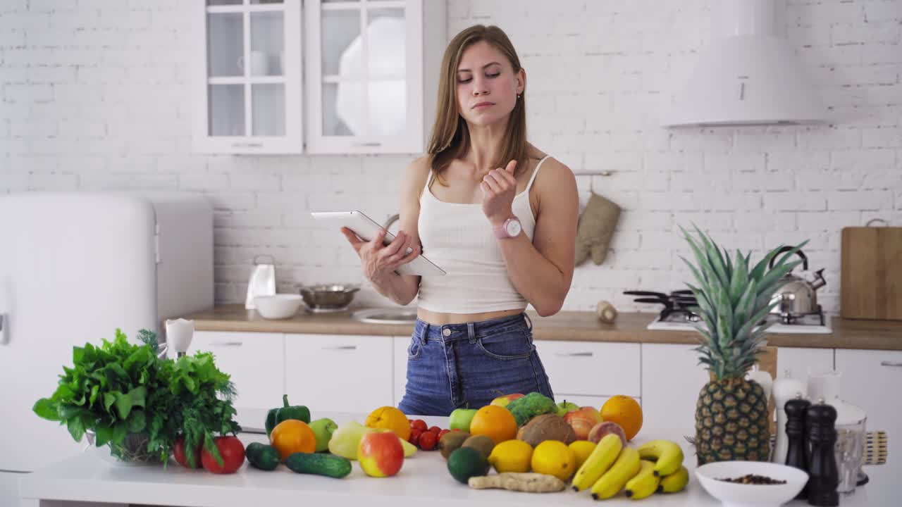 Beautiful woman looking into tablet on the light kitchen background. Young housewife chooses necessary organic fruit and vegetables for recipe to lose weight.