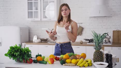 beautiful woman looking into tablet on the light kitchen background. young housewife chooses necessary organic fruit and vegetables for recipe to lose weight.