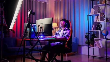 asian boy streamer playing game over network on personal computer, waving hand talking to the participants on the microphone.  live stream video game, desk illuminated by rgb led strip light