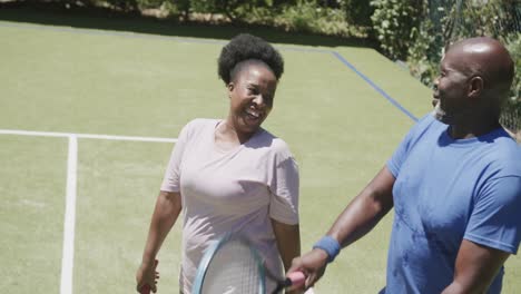 Glückliches-älteres-Afroamerikanisches-Paar,-Das-Mit-Tennisschlägern-Spaziert-Und-Sich-Auf-Dem-Tennisplatz-Unterhält