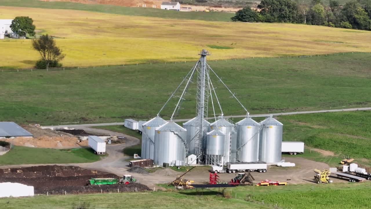 Aerial view of grain storage facility with metal silos in rural Pennsylvania farmland on sunny autumn day. Agricultural infrastructure surrounded by fields and rolling hills. Wide shot