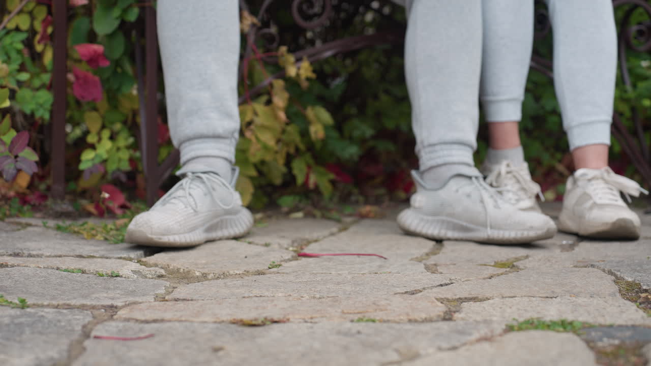 Leg view of two people seated outdoors in matching outfits with light sneakers, standing on stone pavement surrounded by greenery and autumn leaves swaying gently