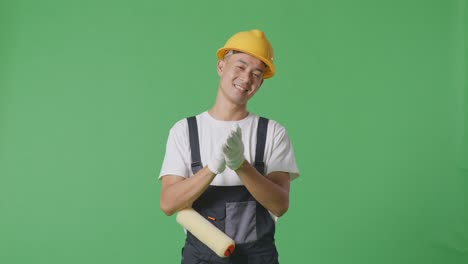 Asian Man Painter Wearing Safety Helmet Smiling And Clapping His Hands While Standing In The Green Screen Background Studio