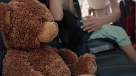 teddy bear in the foreground and mother securing daughter into baby car seat in the background.