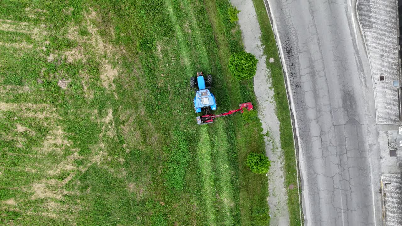 Drone aerial shot showing a blue tractor equipped with a side mower trimming grass along a roadside, leaving visible cutting patterns on the field and maintaining green vegetation