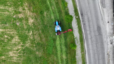drone aerial shot showing a blue tractor equipped with a side mower trimming grass along a roadside, leaving visible cutting patterns on the field and maintaining green vegetation