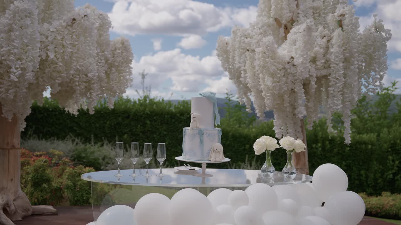 파란 하늘 아래 꽃이 흐드러지게 핀 나무 아래 흰 꽃, 유리 제품, 풍선으로 둘러싸인 층층 케이크
