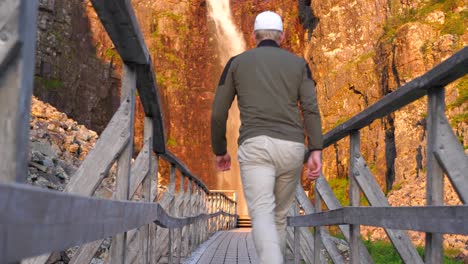 Low-angle-wide-shot-of-man-walking-on-wooden-passageway-towards-Njupeskärs,-Sweden's-highest-waterfall-lit-by-midsummer-morning-golden-sun-in-Fulufjällets-nationalpark