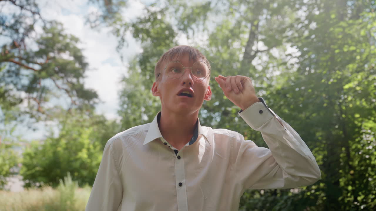 Tall man in white shirt adjusting glasses while carefully observing trees in lush green forest, enjoying peaceful outdoor walk under sunlight surrounded by natural environment and fresh summer