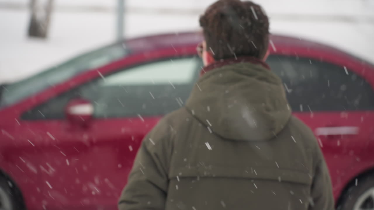 Back view of boy wearing winter jacket walking toward parked car during snow fall with trees in background and snow covering ground on cold outdoor urban day before entering vehicle