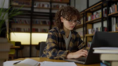 Una-Chica-Segura-De-Sí-Misma,-Con-El-Pelo-Rizado,-Con-Una-Camisa-A-Cuadros-Y-Gafas,-Trabaja-En-Una-Mesa-Usando-Una-Computadora-Portátil-En-Una-Biblioteca-Universitaria