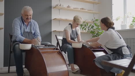 medium shot of middle aged ceramic artist teaching group elderly caucasian woman and senior man how to wedge clay sitting at desk in art studio. people enjoying talking at work