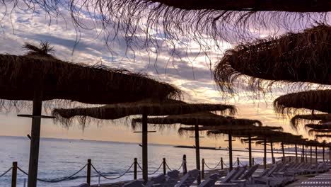 Puesta-De-Sol-De-4k-En-La-Playa-Paradisíaca-De-Marbella,-Hermosas-Nubes-De-Color-Rosa-Púrpura-En-Movimiento-Y-Vista-De-Hamacas-Y-Sombrillas-De-Paja-Tropical