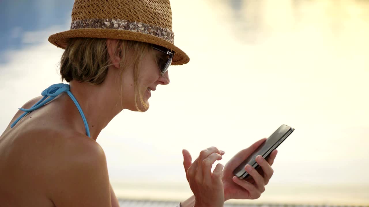 mujer en traje de baño sentada junto a la piscina surfando en teléfono inteligente