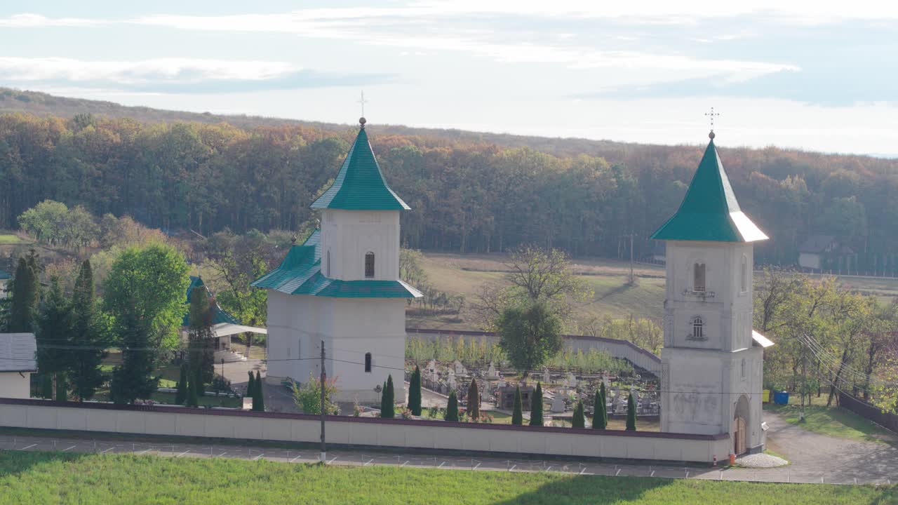 A cinematic aerial shot slowly pushes in towards a traditional Romanian Orthodox monastery, focusing on the church and bell tower with their distinctive green spires and a cemetery. Runc, Buhusi