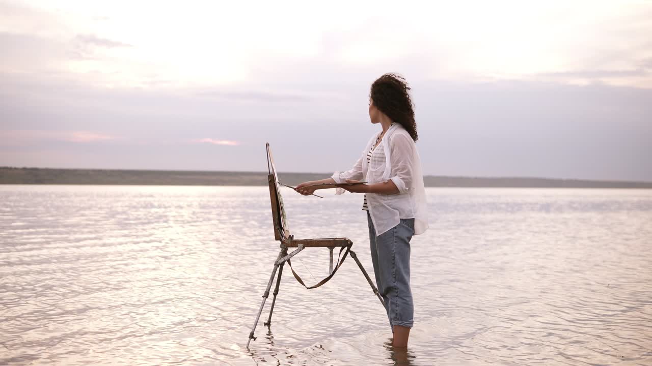 vista deslumbrante de uma jovem morena encaracolada de pé na água com cavalete e desenhando a paisagem circundante. vestindo jeans azuis e camisa branca. comprimento completo