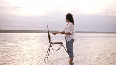 gorgeous view of a young curly brunette standing in the water with easel and draing her the surrounding landscape. wearing a blue jeans and white shirt. full length