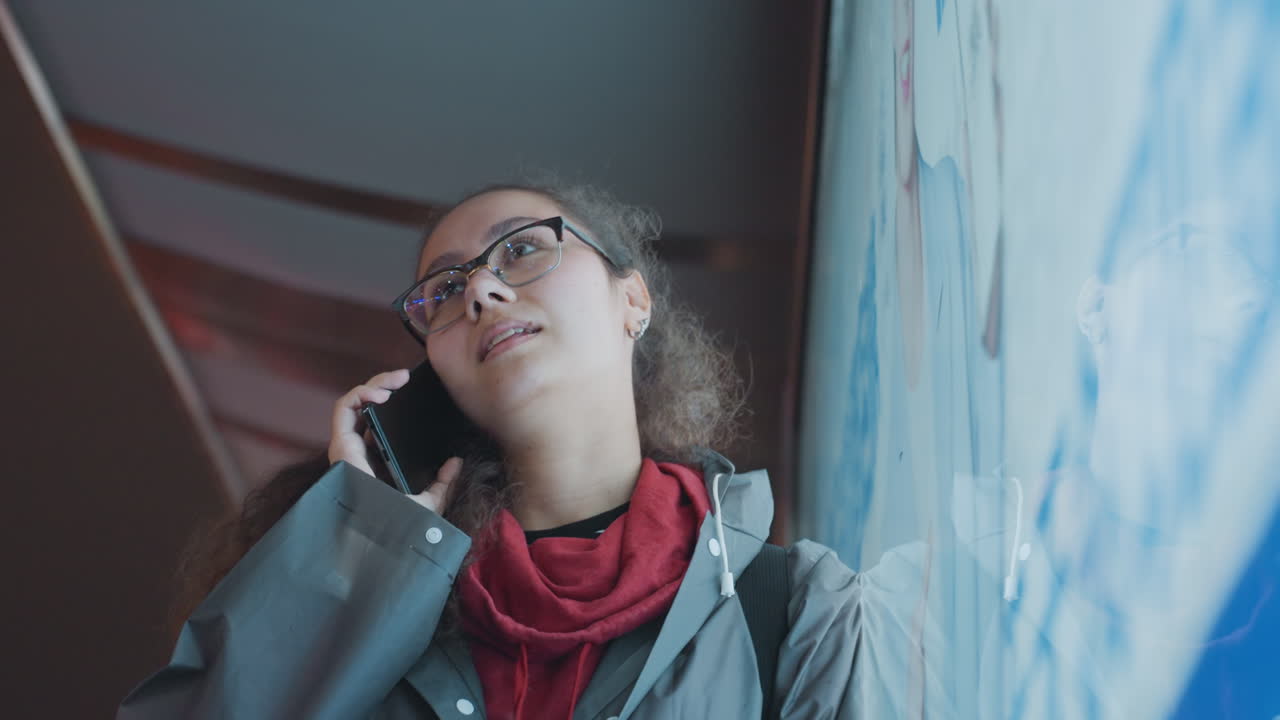Giovane studente birazziale che parla al telefono vicino a un display illuminato, indossa un cappotto grigio e una sciarpa rossa, occhiali, appoggiato a una parete pubblicitaria, con un tenue bagliore blu, superficie riflettente, espressione pensierosa.