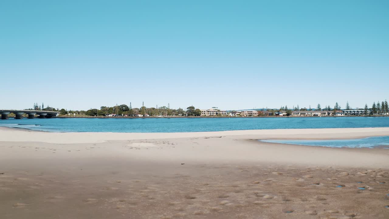 Serene estuary scene with a sandy beach leading to a bridge, overlooking a coastal town in the distance. The peaceful water and blue sky enhance the scenic view