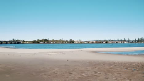 serene estuary scene with a sandy beach leading to a bridge, overlooking a coastal town in the distance. the peaceful water and blue sky enhance the scenic view