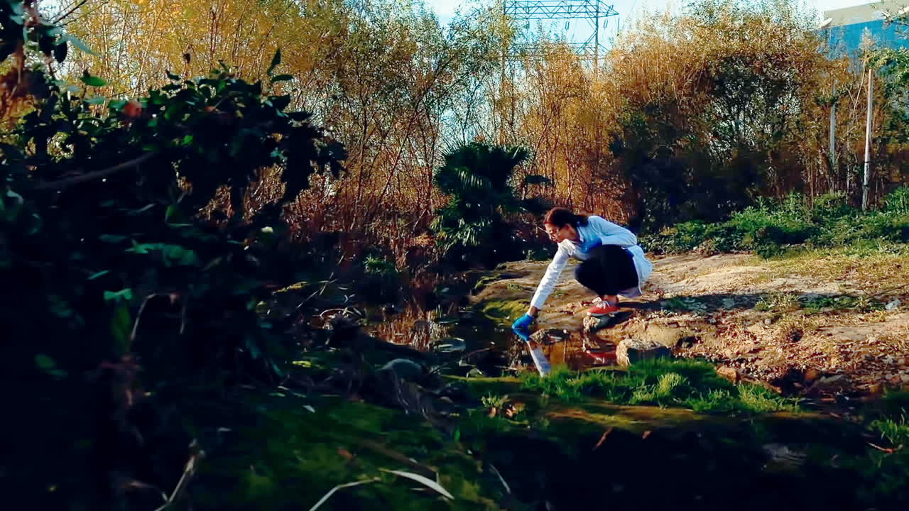 보호 안경과 실험실 코트를 입은 젊은 여성 과학자가 하천에서 물  ⁇ 플을 가져와 관찰하기 위해 들고 있습니다.