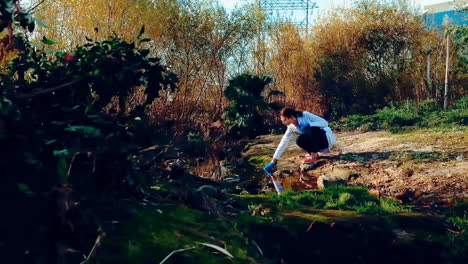 una joven científica en un arroyo, con gafas de protección y una bata de laboratorio, tomando una muestra de agua del arroyo y sosteniéndola para observarla