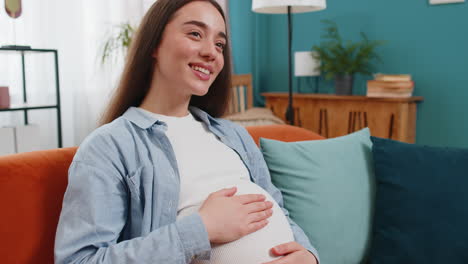 dreamy smiling happy young pregnant woman sitting on sofa at home stroking belly expecting baby