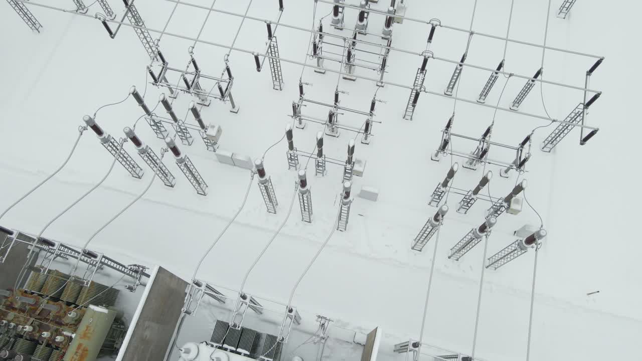 Aerial tilt reveals a frozen electrical substation with visible power transformers, insulators, and grid components resting on pristine snow during an extreme winter day