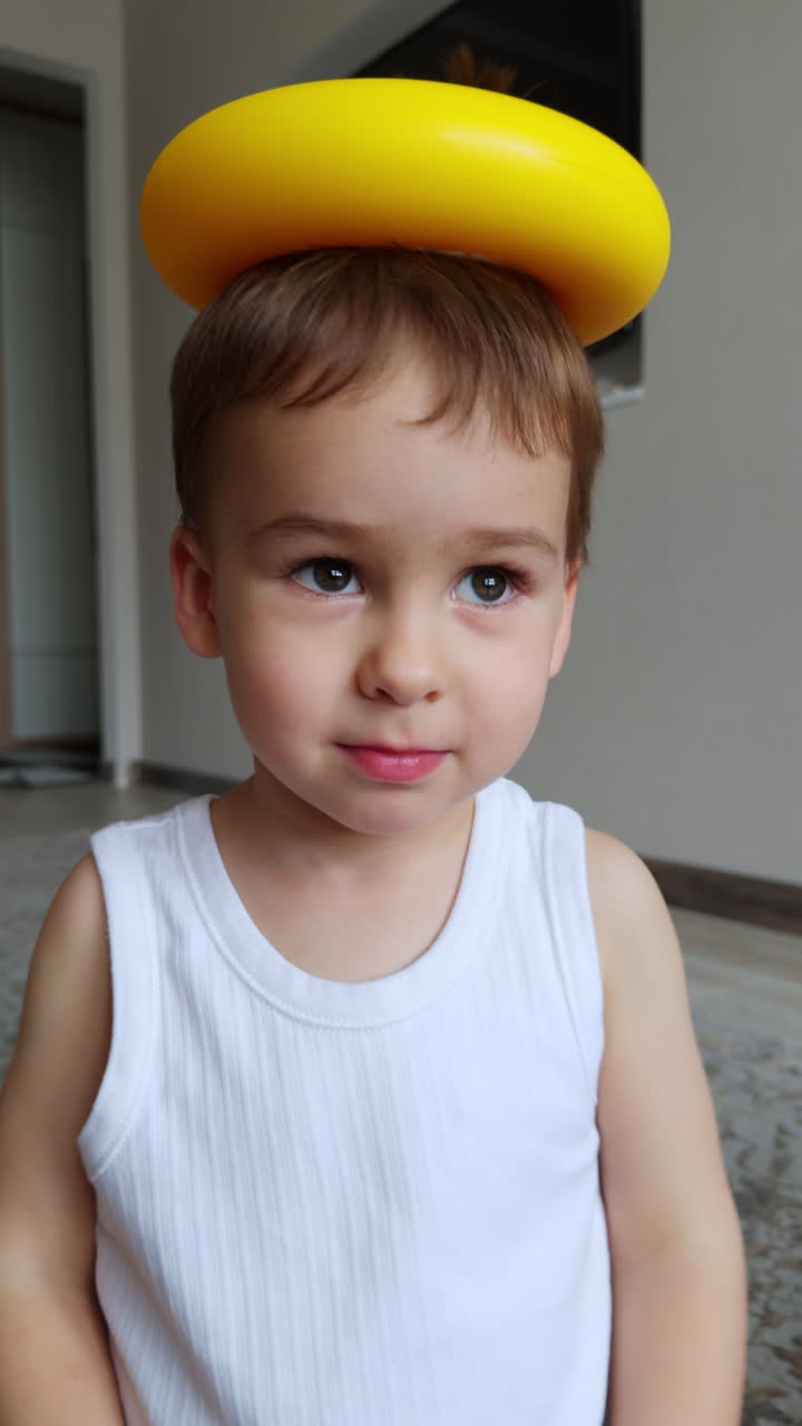 Adorable charming baby boy sits calm holding a toy ring on the head. The toy drops and kid smiles. Close up.