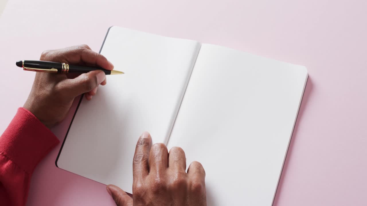 Close up of hands holding pen and blank pages of book, copy space on pink background, slow motion