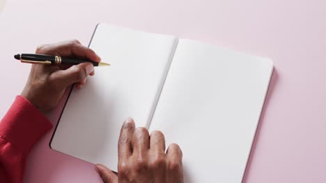 Close-up-of-hands-holding-pen-and-blank-pages-of-book,-copy-space-on-pink-background,-slow-motion