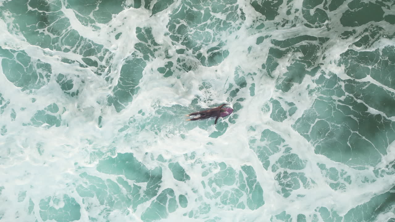 Aerial top down shot of a surfer paddling through thick white foam and over a swell of an incoming wave as he heads back out to the backline, Suances, Spain