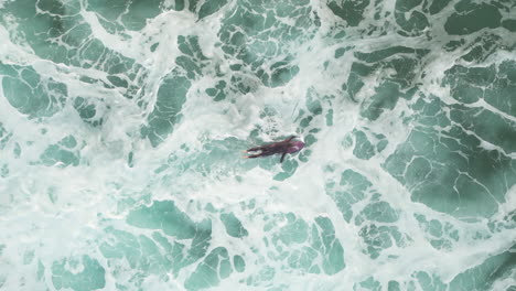 toma aérea de arriba hacia abajo de un surfista remando a través de una espesa espuma blanca y sobre el oleaje de una ola entrante mientras regresa a la línea de fondo, suances, españa