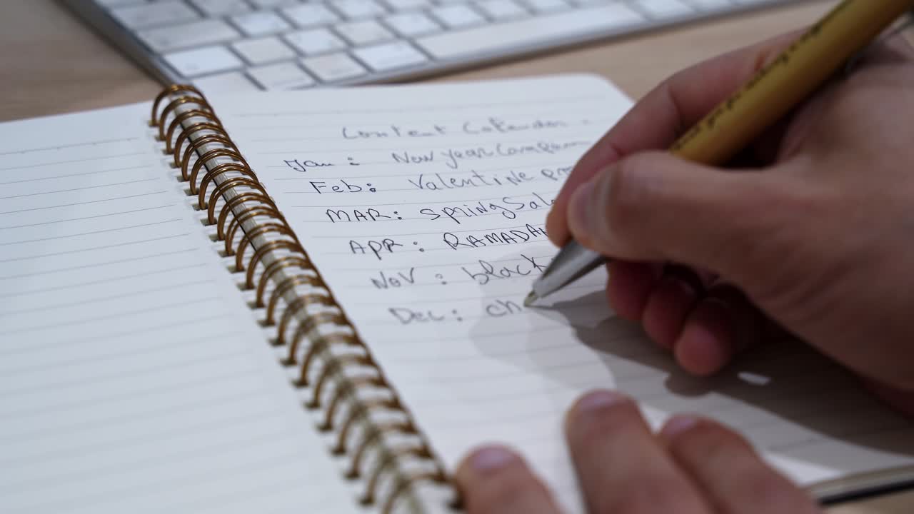 hand actively writing a content calendar with monthly campaigns including Ramadan, Black Friday, and Christmas in a gold spiral notebook on a wooden office desk