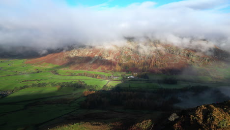 flying over sunlit mountainside towards green valley and mountains shrouded in cloud lit by early morning autumn sunshine