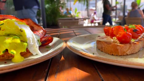 Delicioso-Plato-De-Desayuno-De-Huevos-Benedictinos-Con-Tocino-Crujiente,-Salsa-Holandesa-Y-Aguacate-En-Croquetas-De-Patata-Dulce,-Tostadas-De-Bruschetta-Con-Tomates-Cherry,-Huevos-Escalfados-Para-El-Brunch-En-Un-Restaurante,-4k