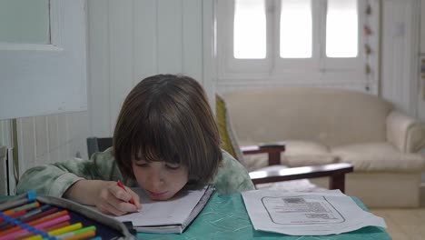 close view of a boy writing on an exercise book with his chin on the table,
