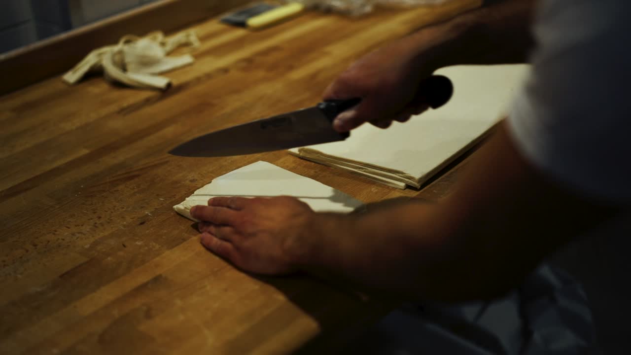 Nærbillede i slow motion af en mand, der stabler lamineret dej i bogstavfold, i croissantfremstillingsprocessen.