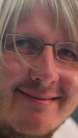una vista facial de un científico sonriente con una peluca rubia y gafas, sentado en un laboratorio con un microscopio con reflejo de luz visto en su cabeza