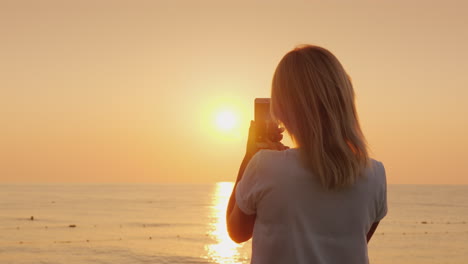 Eine-Junge-Blonde-Frau-Fotografiert-Auf-Einem-Smartphone-Eine-Rosa-Morgendämmerung-Am-Meerblick-Von-Hinten