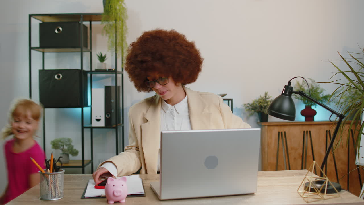 Tired businesswoman working on laptop at home office, hyperactive child girl running, making chaos