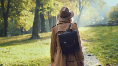 rear view of caucasian woman in coat, hat and with a backpack walking in the park and looking at the sides in autumn