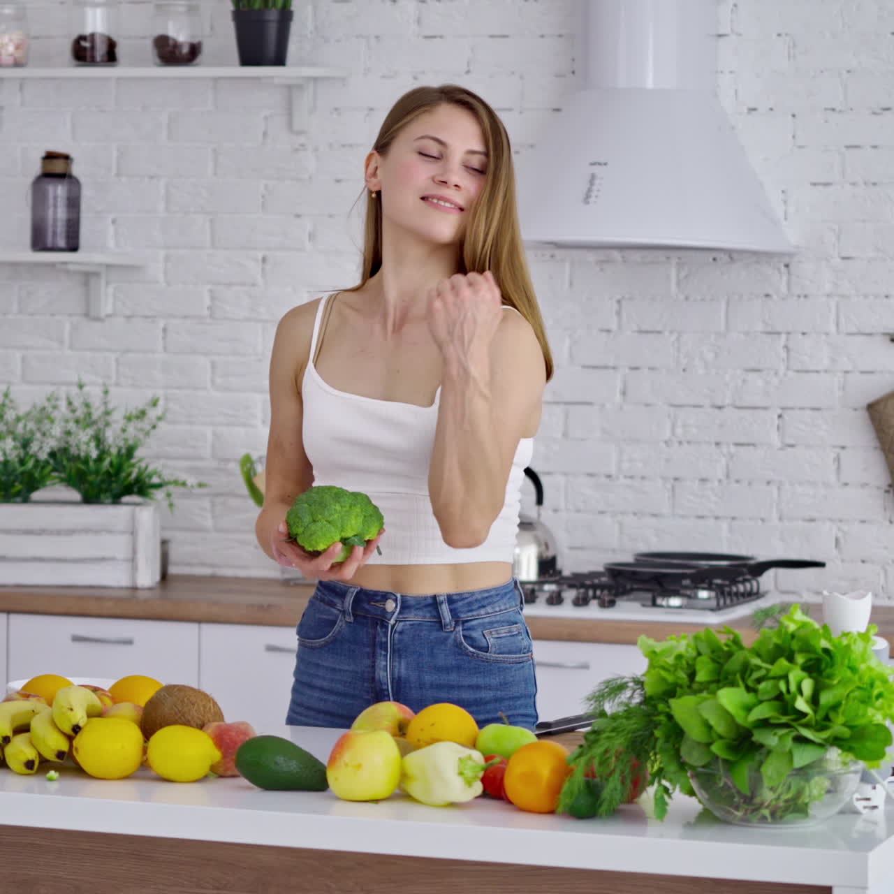 Attractive woman at the table with fresh vitamins. Beautiful sportswoman in casual wearing holding broccoli and shows her arm muscles in the kitchen. Healthy lifestyle.