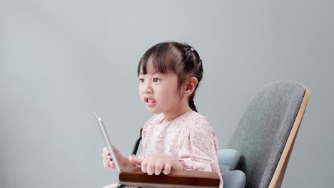 asian child with fringe in hair sitting on chair laughing while using digital tablet