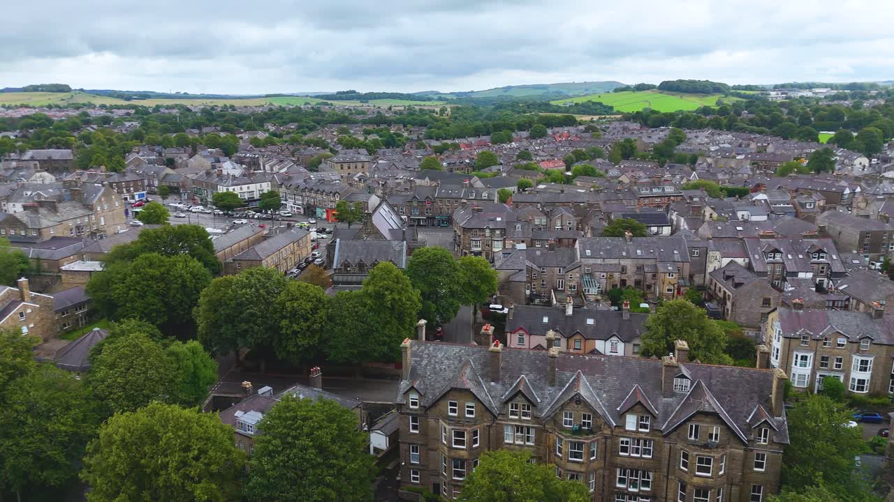 Drone camera smoothly pans above Buxton, England, revealing historic stone buildings, tree-lined streets, and rolling green hills under overcast daylight